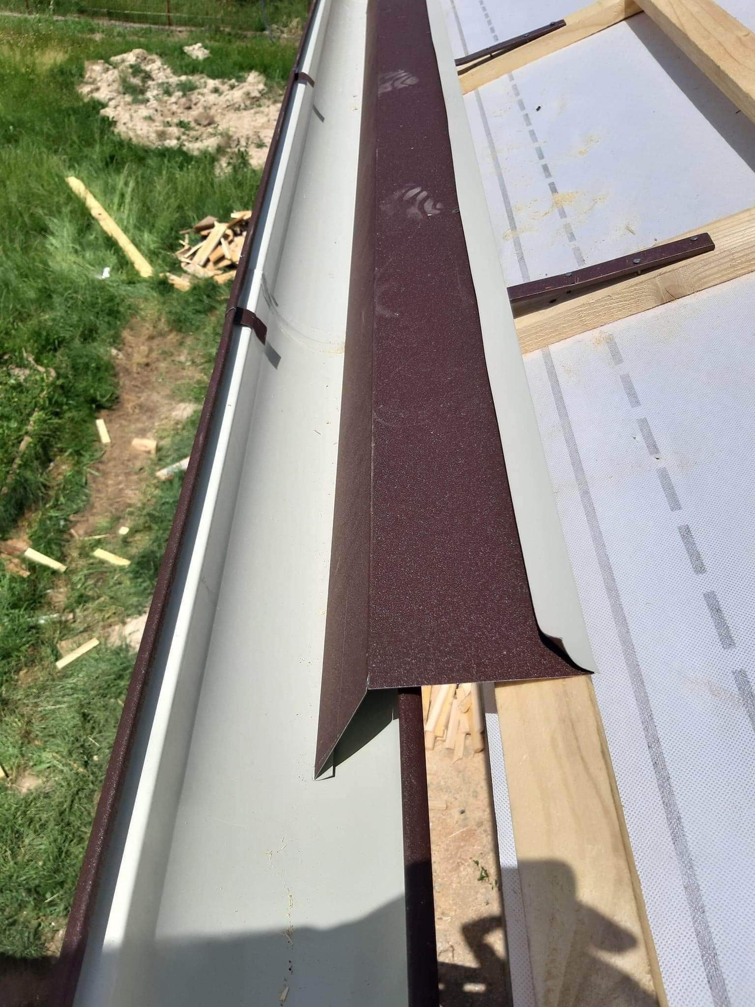 Szara rynna z brązowym narożnikiem zamontowana na drewnianej konstrukcji dachu, widok z góry, w tle trawa i resztki drewna.