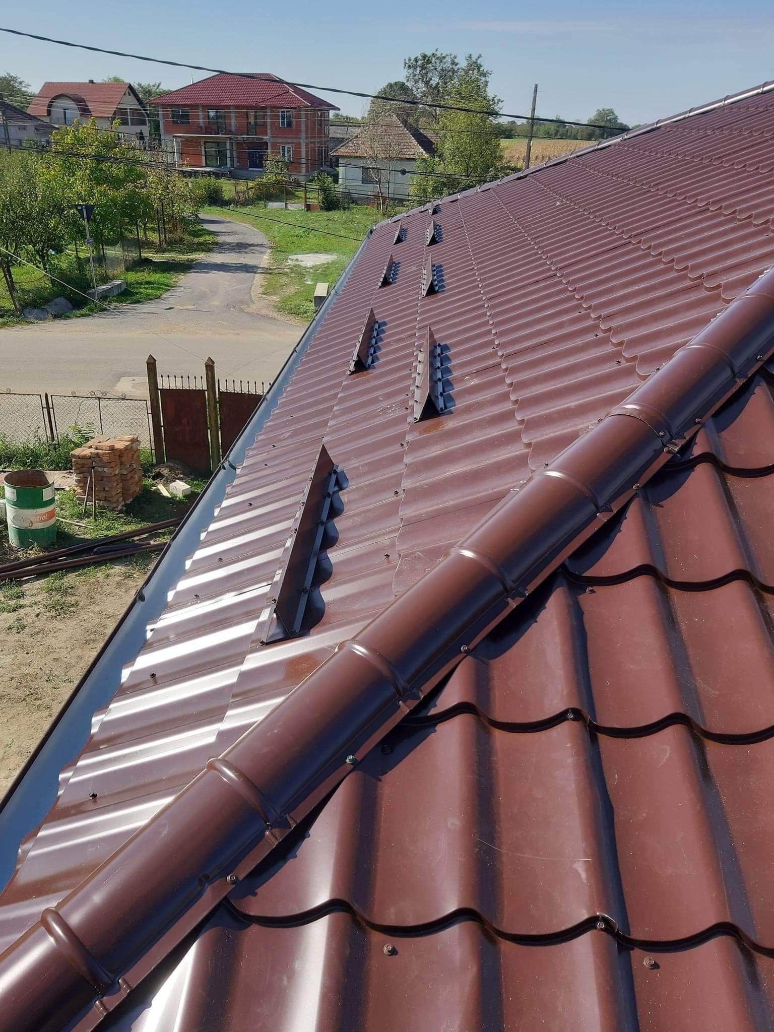 Ciemnobrązowy dach z blachy trapezowej z widocznymi kominkami wentylacyjnymi i elementami odprowadzającymi wodę, w tle zabudowa jednorodzinna i błękitne niebo.