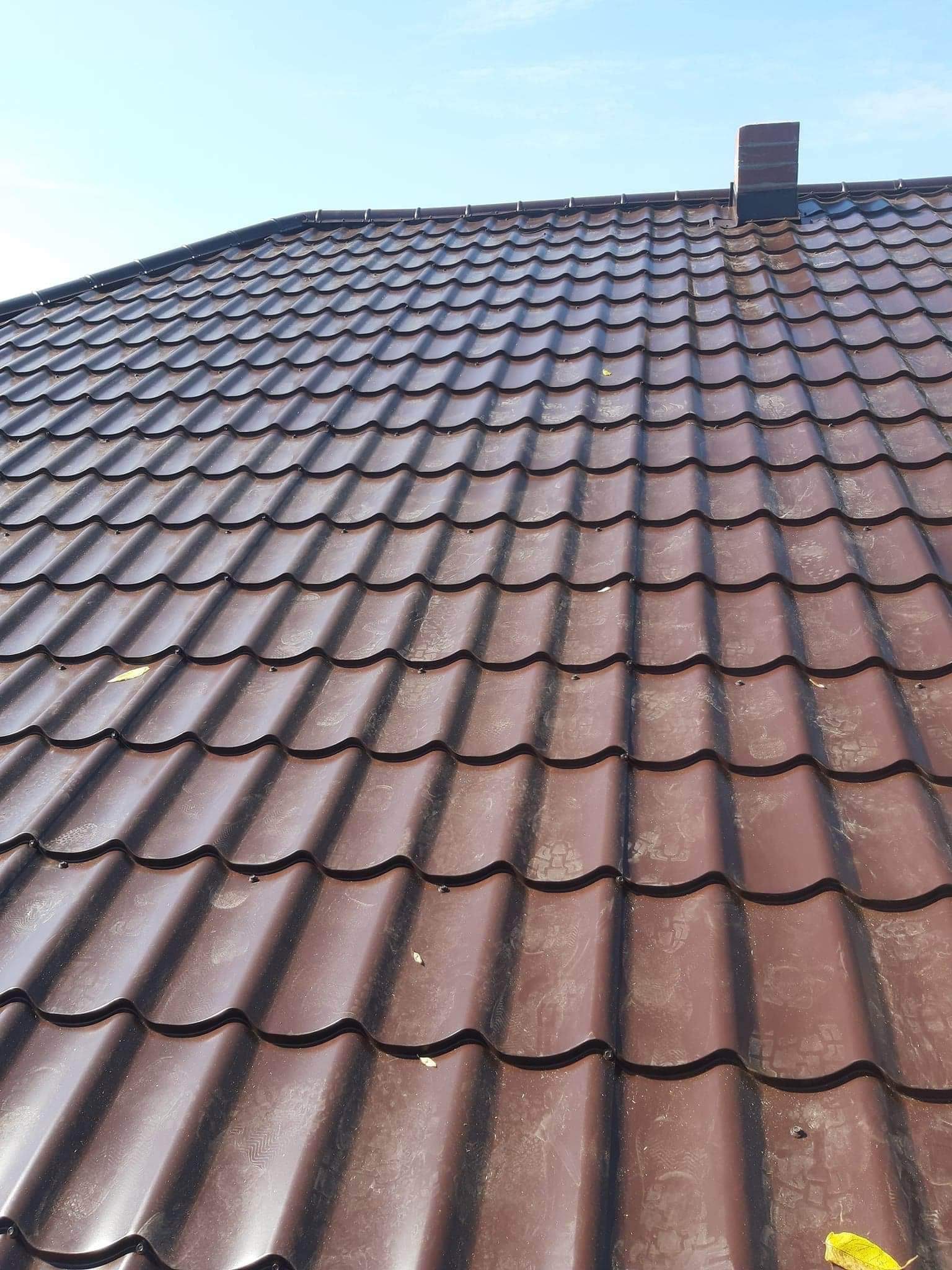 Widok z góry na brązowy dach z blachodachówki modułowej, z kominem w prawym górnym rogu i pojedynczymi liśćmi na powierzchni.