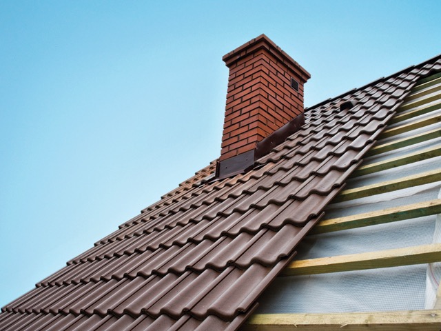 Brunatny dach z dachówki ceramicznej, częściowo w trakcie montażu z widoczną drewnianą konstrukcją więźby i kominem z czerwonej cegły na tle błękitnego nieba.