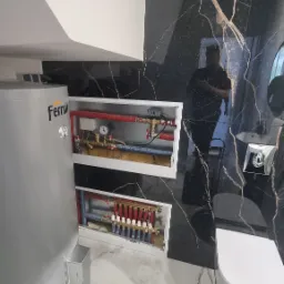 Szary bojler Ferroli w łazience z czarnym marmurem. Widoczne rozdzielacze ogrzewania podłogowego w białych wnękach. Nowoczesne wykończenie.