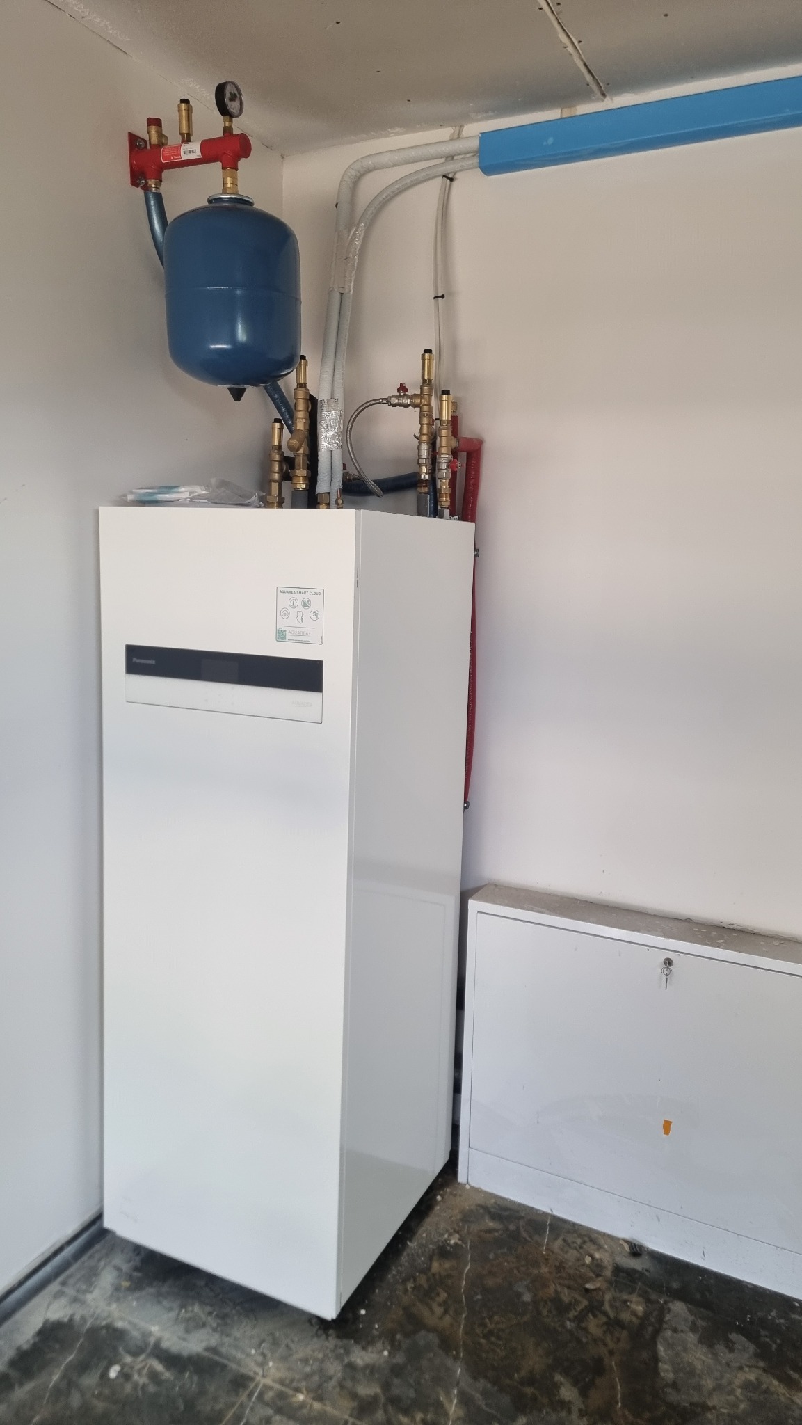 Biała pompa ciepła Panasonic Aquarea z niebieskim zbiornikiem wyrównawczym i instalacją hydrauliczną w pomieszczeniu technicznym. Widoczne rury i zawory.