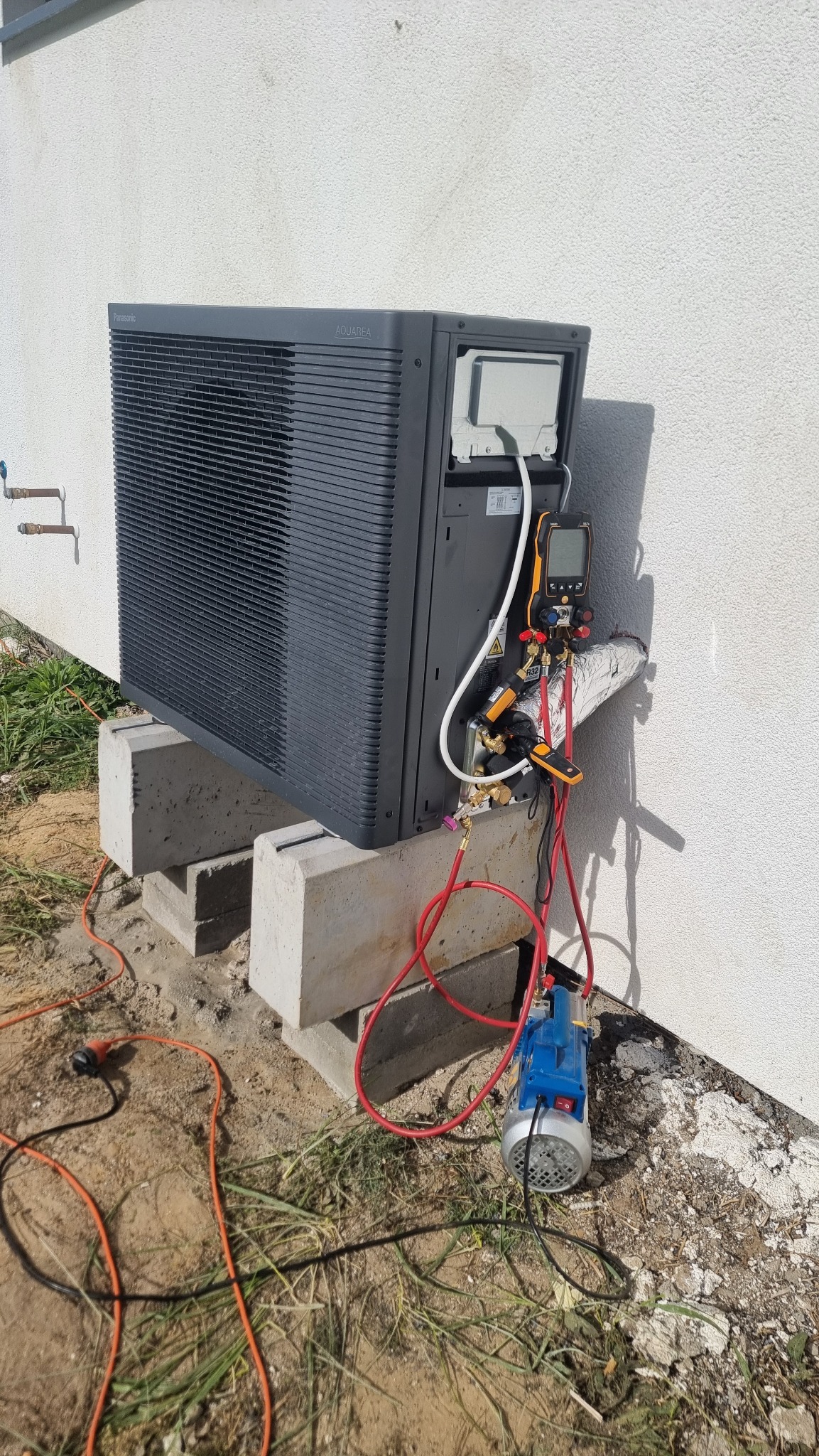 Szara pompa ciepła Panasonic Aquarea zamontowana na betonowych blokach przy białej ścianie budynku, podłączona do narzędzi serwisowych.