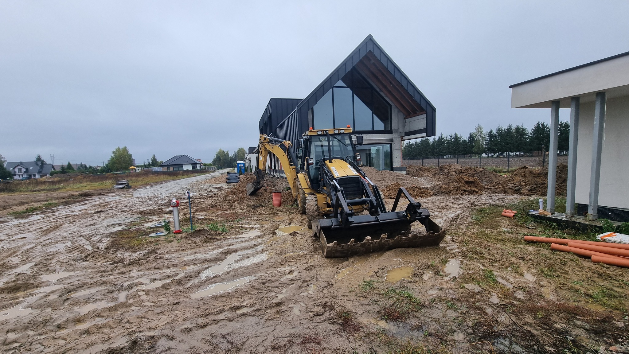 Żółta koparko-ładowarka na błotnistym placu budowy przed nowoczesnym domem z dużymi oknami. Widoczne rury i elementy instalacji. Pochmurne niebo.