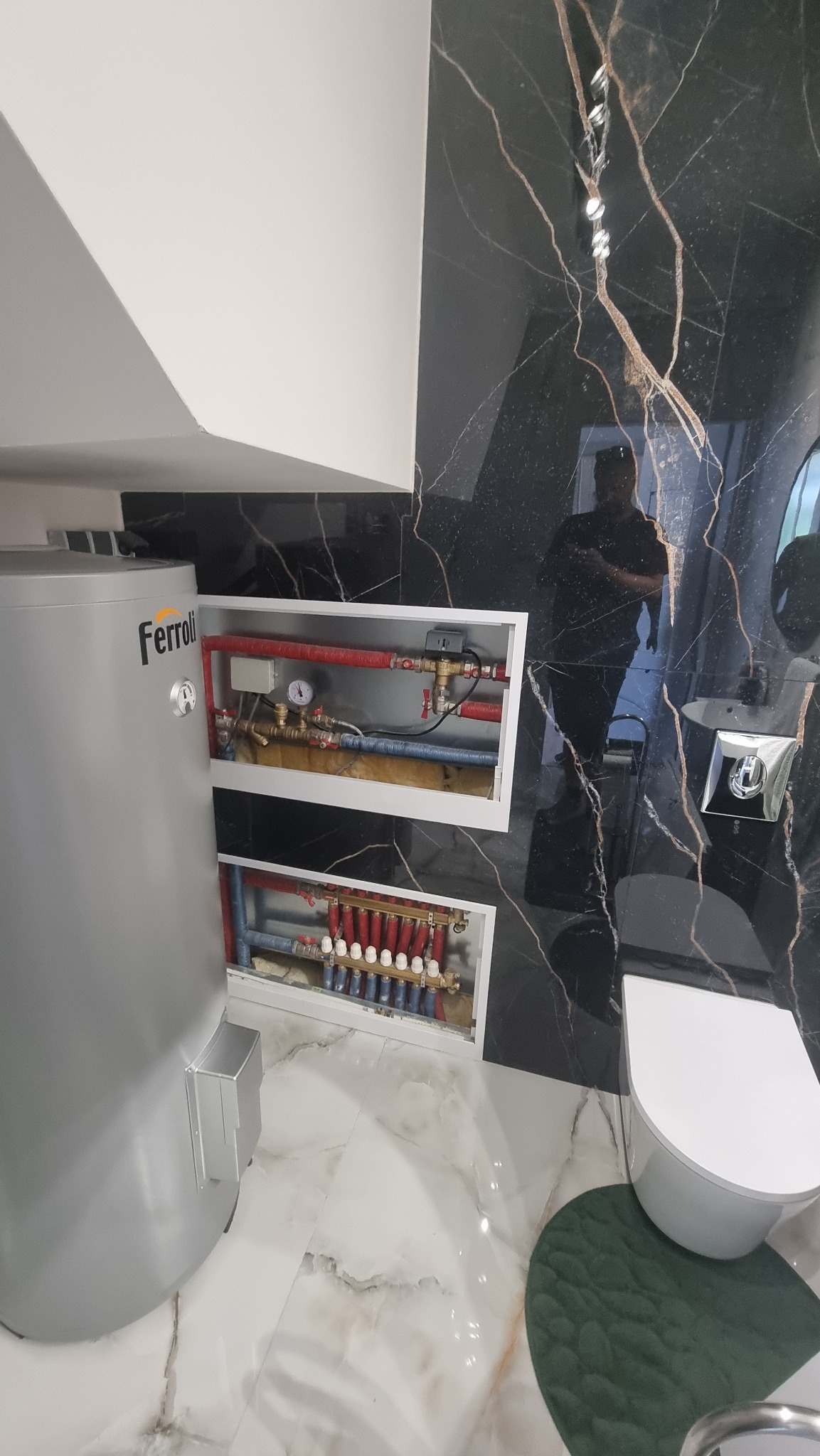 Szary bojler Ferroli w łazience z czarnym marmurem. Widoczne rozdzielacze ogrzewania podłogowego w białych wnękach. Nowoczesne wykończenie.