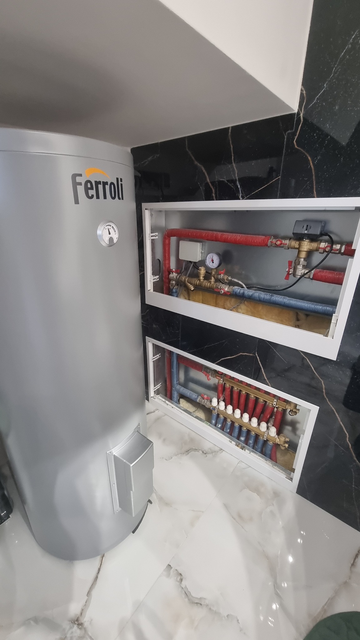 Szary zbiornik Ferroli obok rozdzielacza ogrzewania podłogowego w białej obudowie na tle marmurowej ściany. Widoczne czerwone i niebieskie rury instalacji.