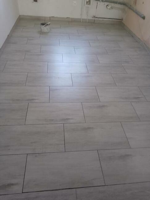 Ułożone, poziome płytki podłogowe w odcieniach szarości, z widocznym wiadrem i pionowymi rurami w tle, prezentujące postęp prac remontowych.