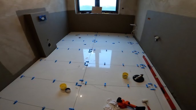 Białe płytki podłogowe w trakcie układania w łazience, z użyciem systemu poziomowania. Widoczne narzędzia i poziomica. Częściowo wykończone ściany w szarym kolorze.