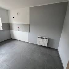 Wnętrze pokoju po częściowym szpachlowaniu ścian na szaro i biało, z grzejnikiem na ścianie i szarą podłogą z płytek. Widok na narożnik pokoju.