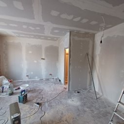Pokój w trakcie remontu: ściany i sufit pokryte płytami karton-gips, widoczne przewody, drabina i narzędzia. Surowy stan wykończenia wnętrza.