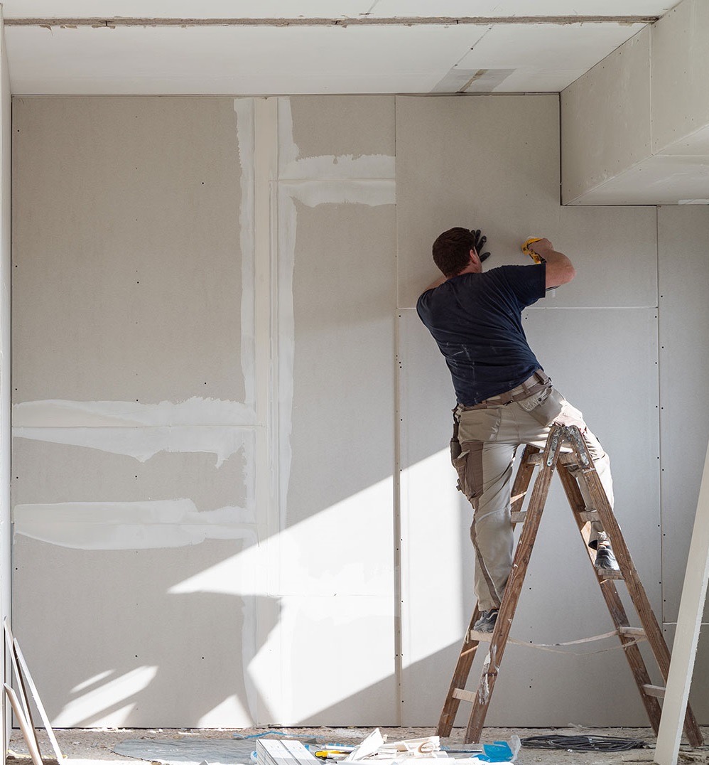 Pracownik na drabinie montuje płytę gipsowo-kartonową na ścianie. Widoczne łączenia płyt i wstępnie zaszpachlowane miejsca. Jasne wnętrze w trakcie remontu.