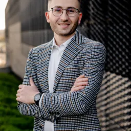 Uśmiechnięty mężczyzna w okularach i marynarce w kratę, stojący z założonymi rękami na tle nowoczesnej fasady budynku z metalowymi elementami.