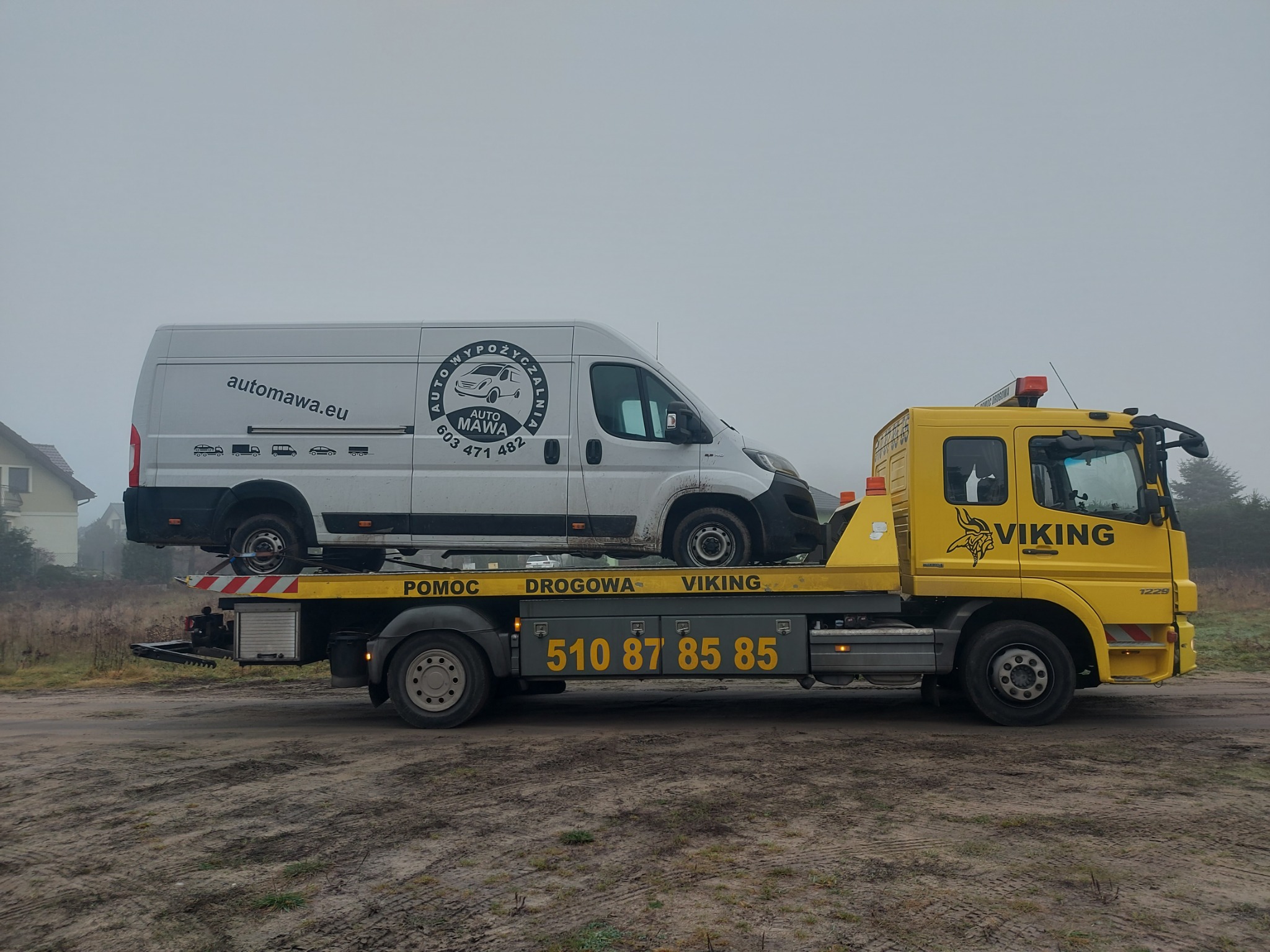 Żółta laweta z logiem Viking przewozi brudny biały bus z reklamą wypożyczalni samochodów Automawa na tle zamglonego krajobrazu.