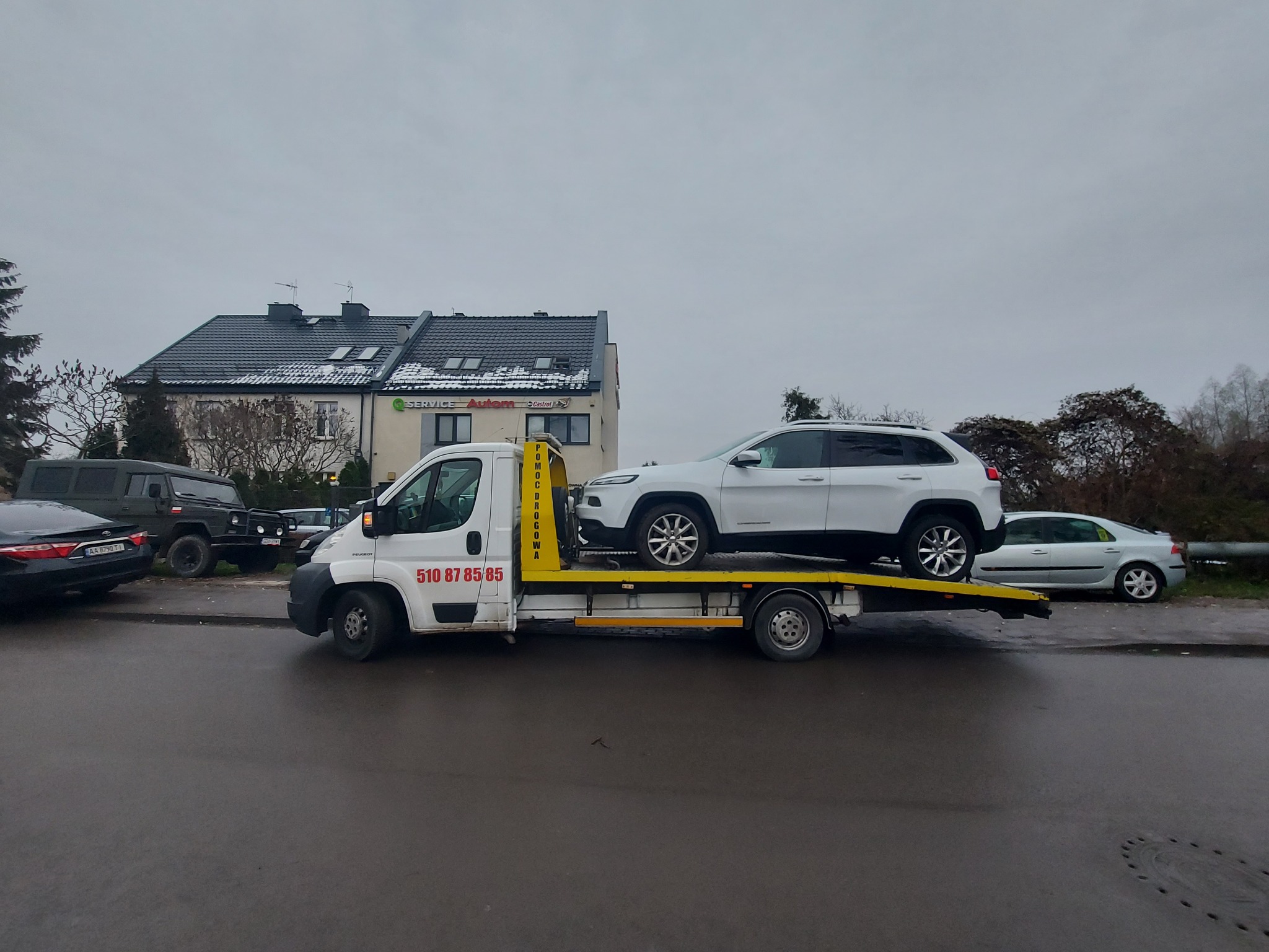 Biała laweta z białym SUV-em na platformie, zaparkowana na mokrej ulicy w pochmurny dzień, z budynkiem usługowym w tle.