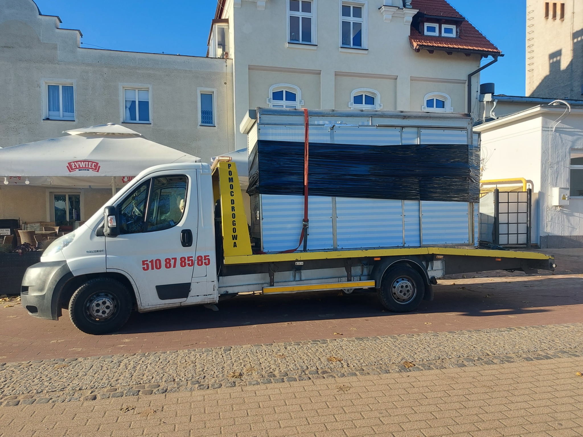 Biała laweta z napisem 'POMOC DROGOWA' na żółtym panelu, przewożąca zabezpieczony czarną folią ładunek w zabudowie kontenerowej, zaparkowana na chodniku z kostki brukowej w tle zabudowań miejskich.