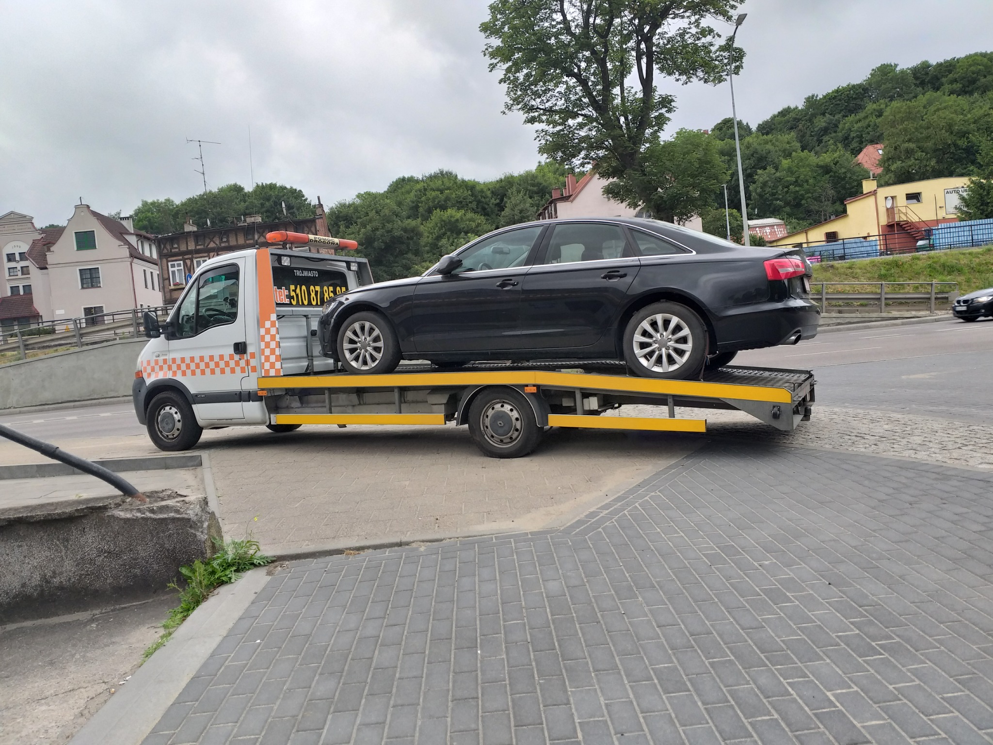 Czarny samochód osobowy marki Audi A6 na żółtej platformie pojazdu pomocy drogowej z pomarańczowymi światłami ostrzegawczymi, zaparkowanego na kostce brukowej w otoczeniu zabudowy miejskiej i zieleni.