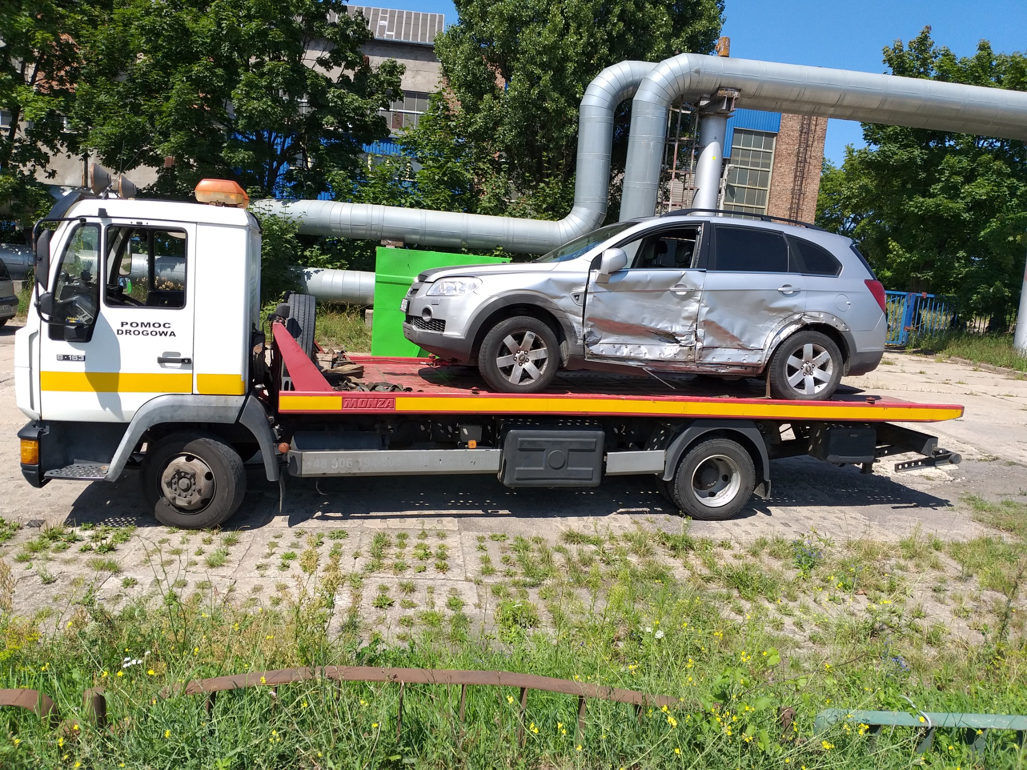 Laweta z uszkodzonym srebrnym SUV-em marki Chevrolet Captiva, widoczne wgniecenia na drzwiach i błotniku, na tle przemysłowego krajobrazu z rurami i zielenią.