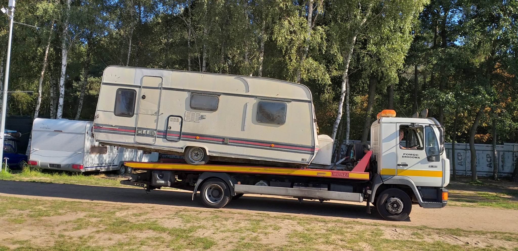 Laweta z białym ciągnikiem przewozi przyczepę kempingową w beżowym kolorze z czerwoną linią na tle drzew i zieleni.