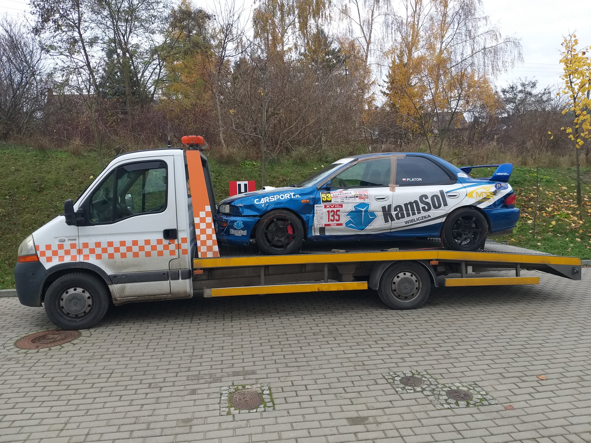 Biała laweta z pomarańczowymi elementami przewozi niebieski samochód rajdowy marki Subaru z naklejkami sponsorskimi na tle drzew i chodnika z kostki brukowej.