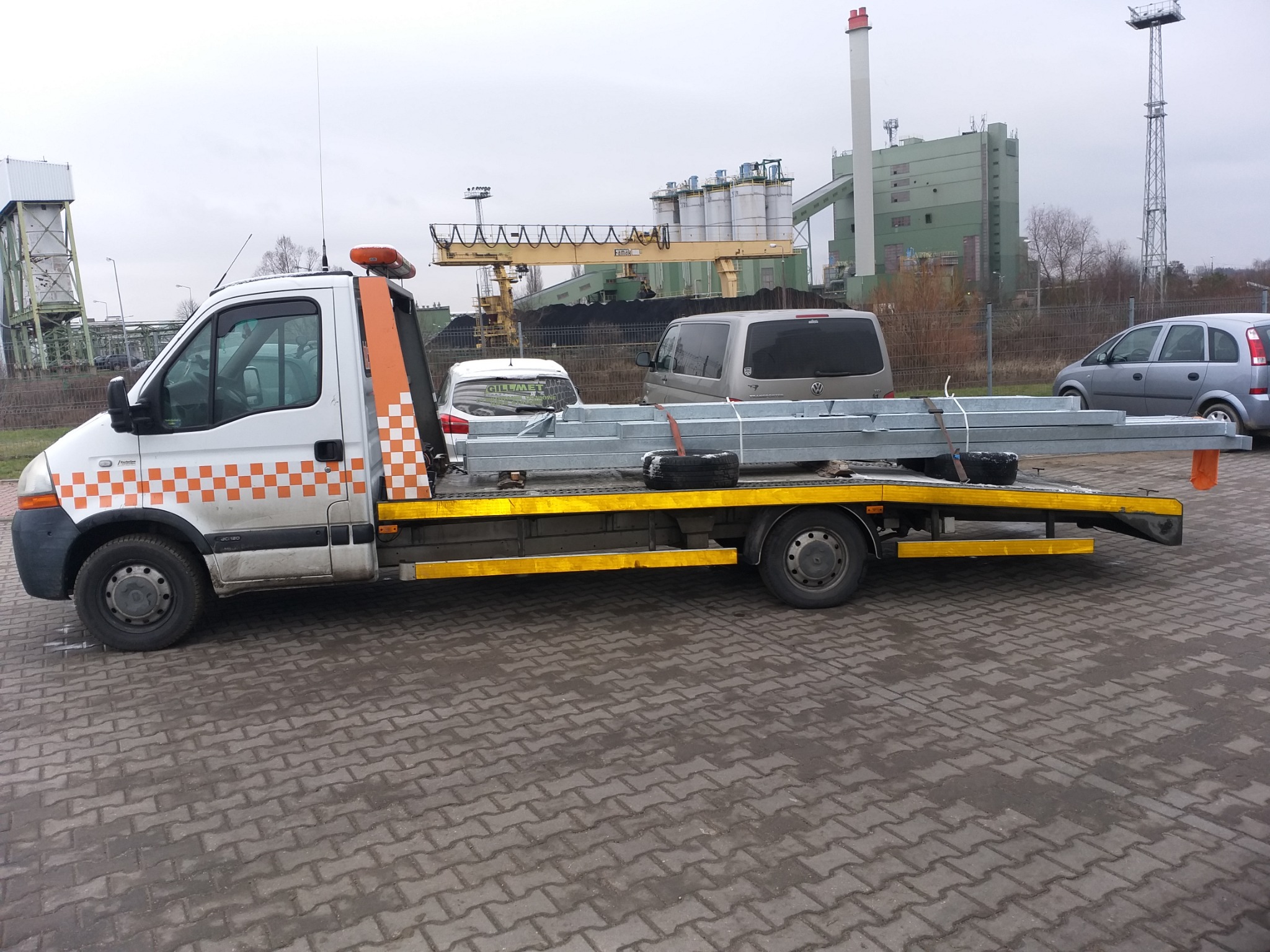 Biała laweta z pomarańczowo-białą szachownicą na kabinie, przewożąca stalowe profile i opony, na tle przemysłowego krajobrazu z dźwigiem i budynkami fabrycznymi.