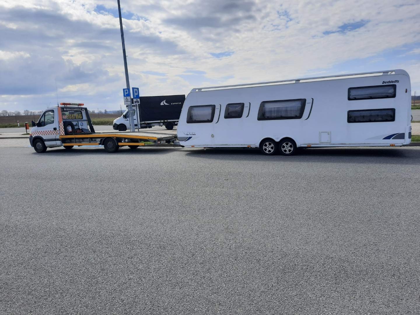 Pomarańczowo-biała autolaweta holuje białą przyczepę kempingową na parkingu, w tle widoczny zaparkowany czarny samochód dostawczy.