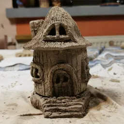 Ręcznie wykonana, miniaturowa gliniana chatka z trzcinowym dachem, imitująca dom dla lalek lub element dekoracyjny, na stole pokrytym płótnem.