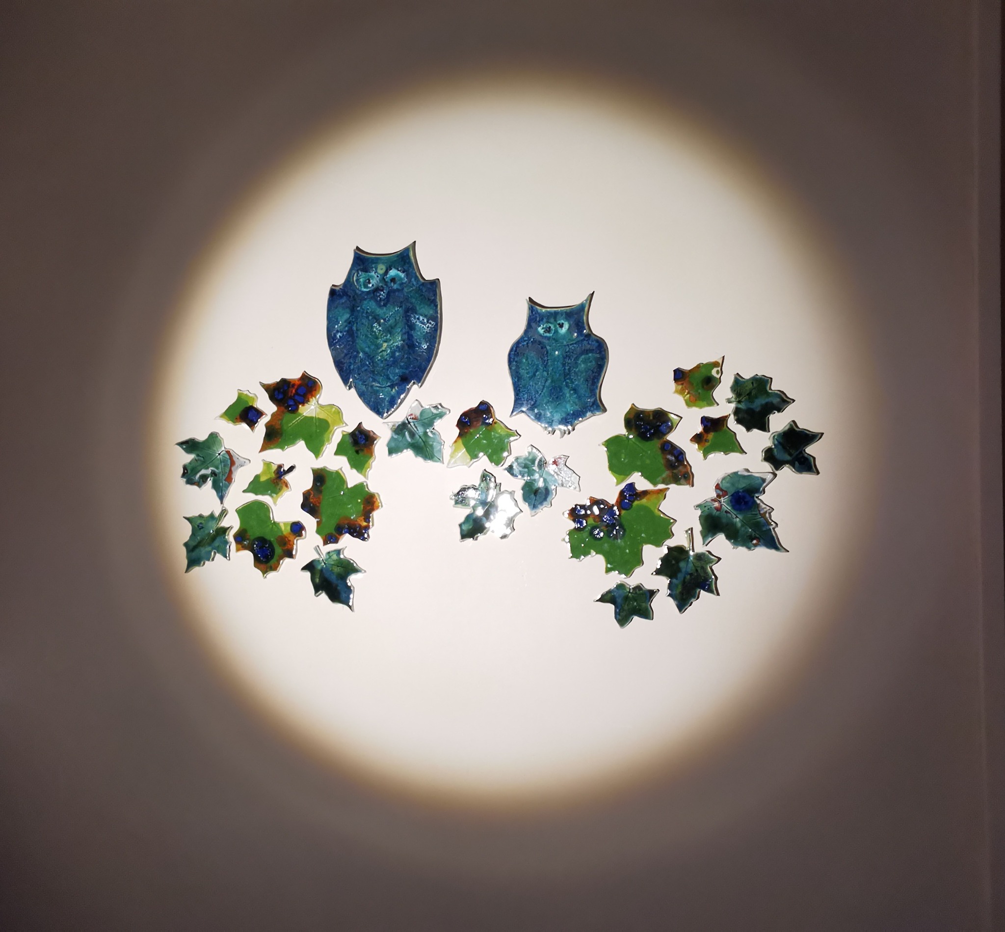 Dekoracja ścienna z emaliowanymi elementami w kształcie sów i liści, oświetlona punktowym światłem, tworząca artystyczną kompozycję.