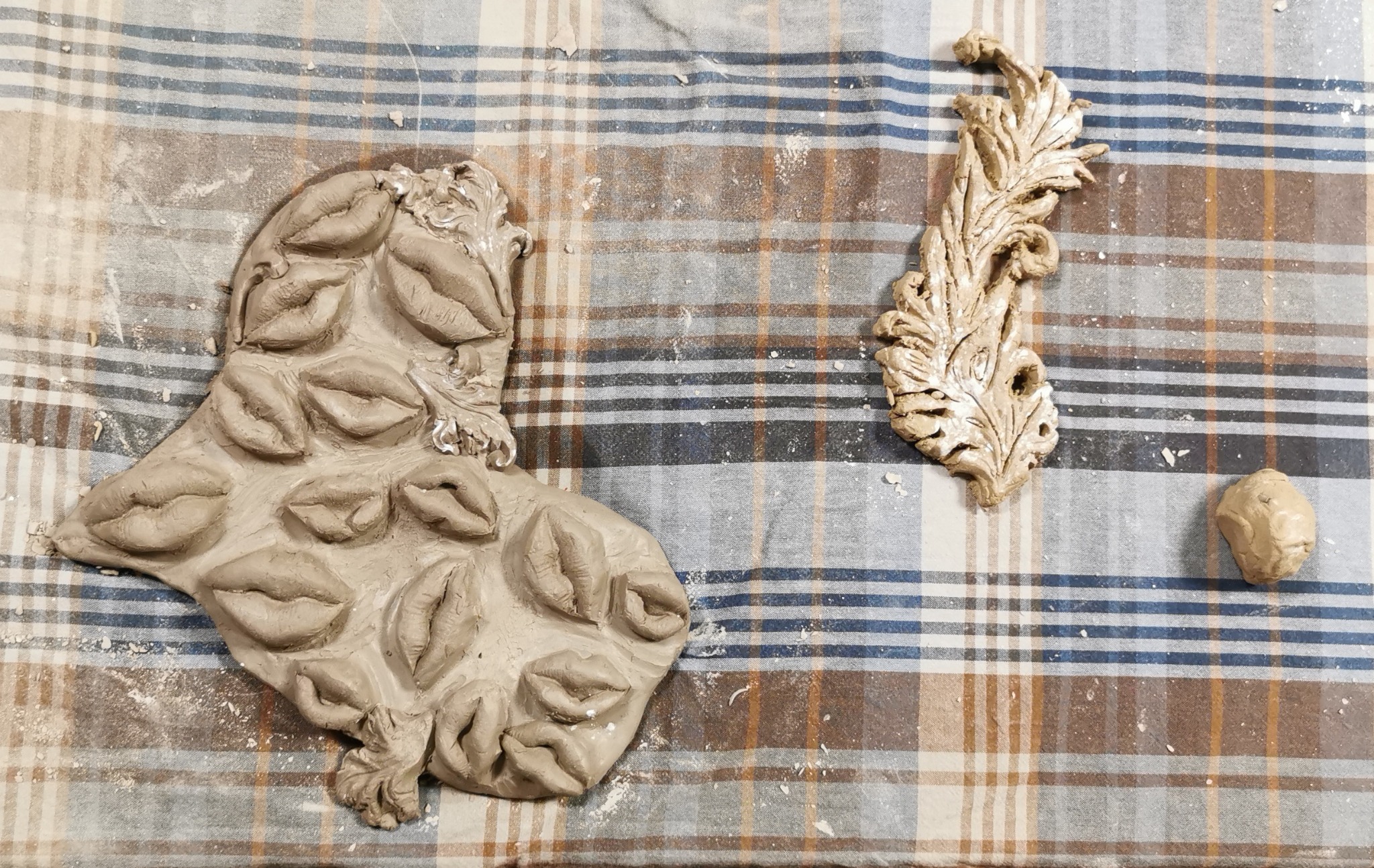Dwie gliniane rzeźby: jedna przedstawia płaskorzeźbę z ustami, druga ornament roślinny, leżące na kraciastej tkaninie z widocznymi okruchami gliny.