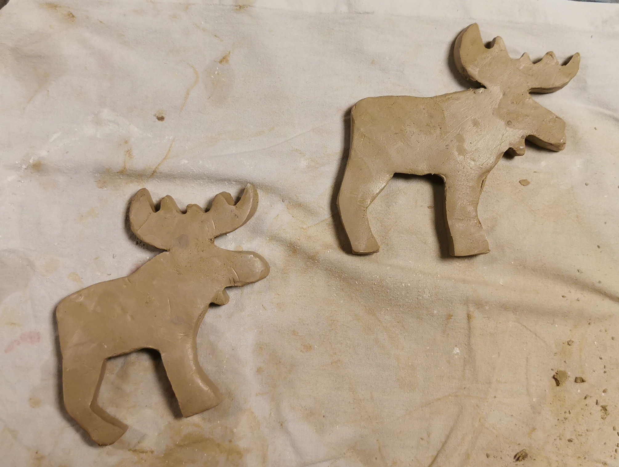Dwa surowe, gliniane modele łosi o nieregularnych krawędziach, leżące na brudnej, kremowej tkaninie z widocznymi okruchami gliny.