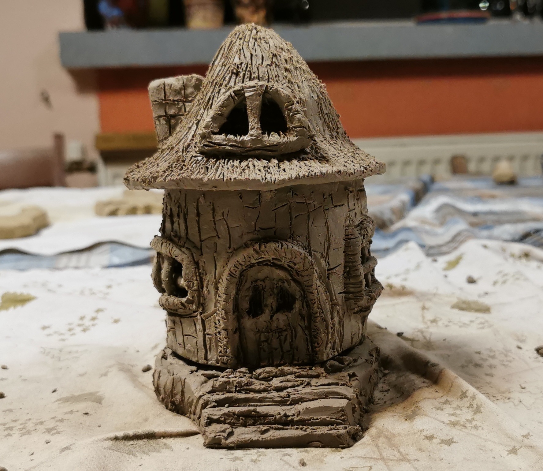 Ręcznie wykonana, miniaturowa gliniana chatka z trzcinowym dachem, imitująca dom dla lalek lub element dekoracyjny, na stole pokrytym płótnem.