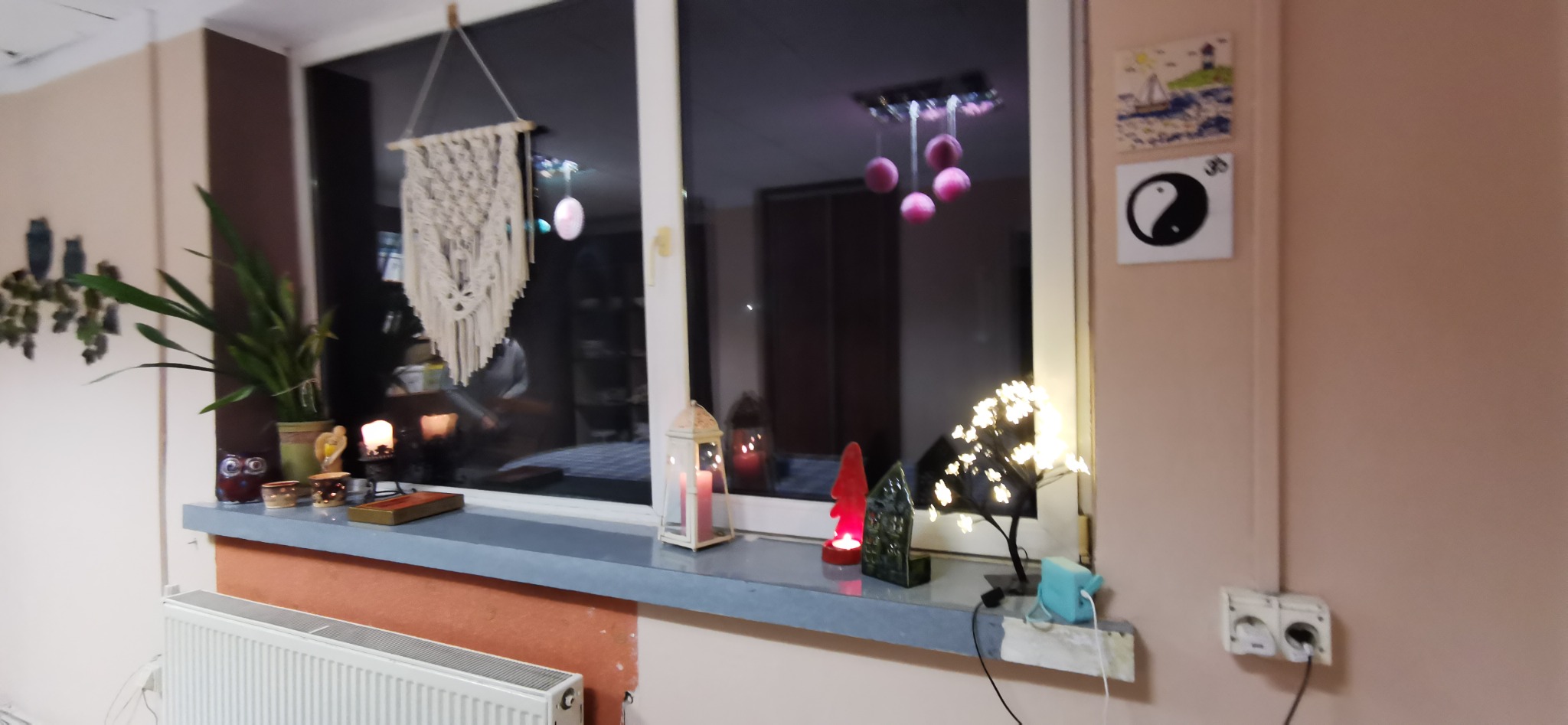 Dekoracyjny parapet z roślinami, świecami w lampionach, makramą i ozdobną lampką w kształcie drzewka z drobnymi światełkami, tworzący przytulną atmosferę.