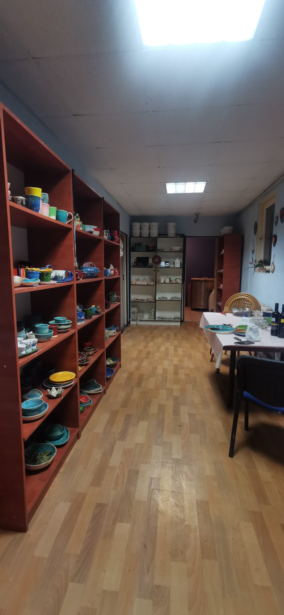 Wnętrze pracowni ceramicznej z regałami pełnymi ręcznie robionych naczyń, stołem przygotowanym do warsztatów i ekspozycją wyrobów w tle.