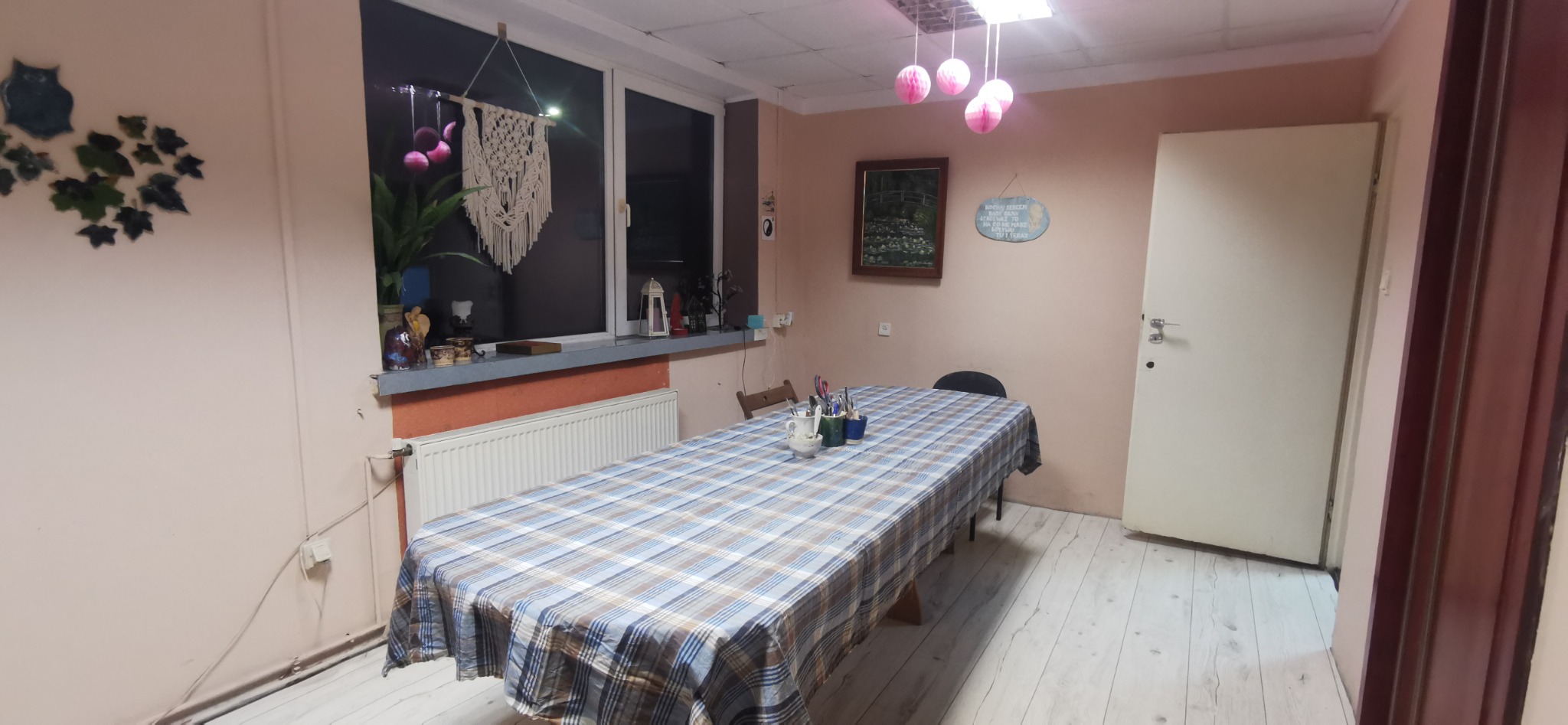 Pokój z długim stołem przykrytym niebiesko-białą kratą, przy oknie rośliny i dekoracje, na ścianie obrazek i ozdobny napis, lampa z różowych kul.