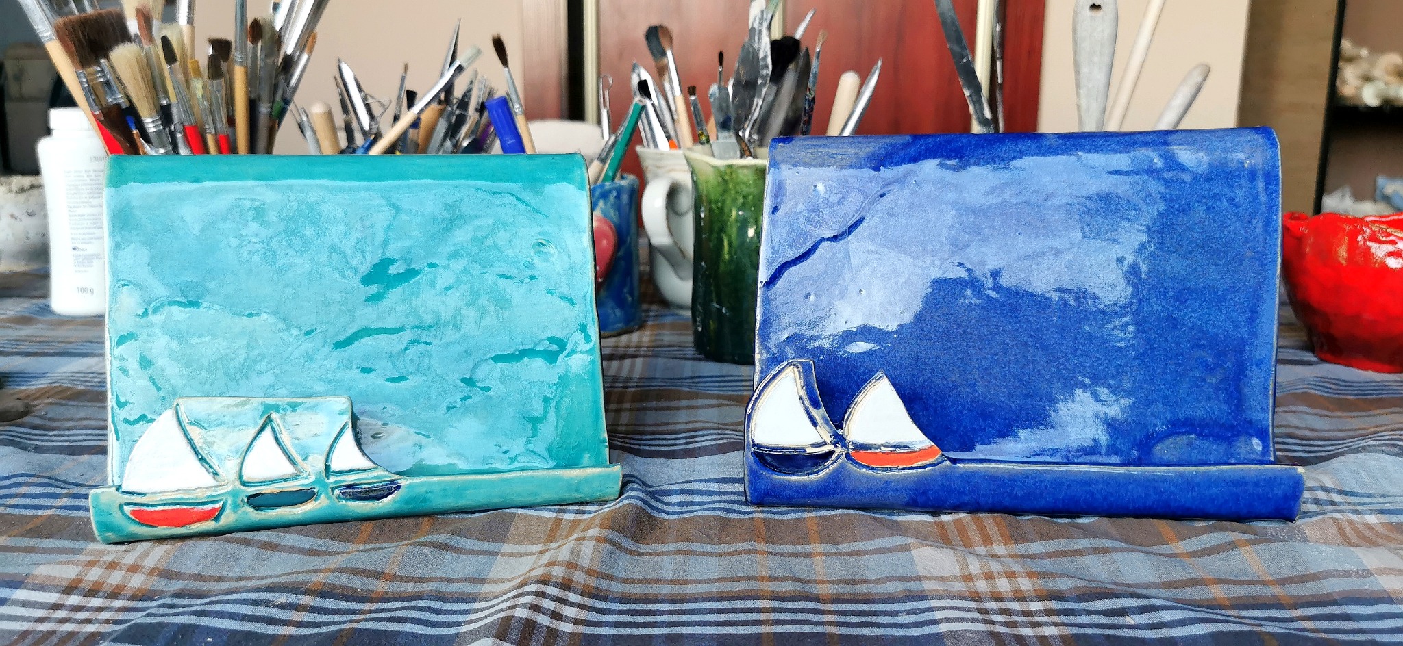 Dwa ceramiczne, ręcznie robione stojaki na książki lub tablety, jeden turkusowy, drugi granatowy, oba z motywem żaglówek, ustawione na kraciastej tkaninie, w tle pędzle i przybory artystyczne.
