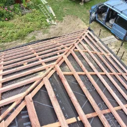 Fragment konstrukcji dachu z drewnianymi łatami na czarnej membranie, widok z góry, w tle niebieski samochód i koparka.