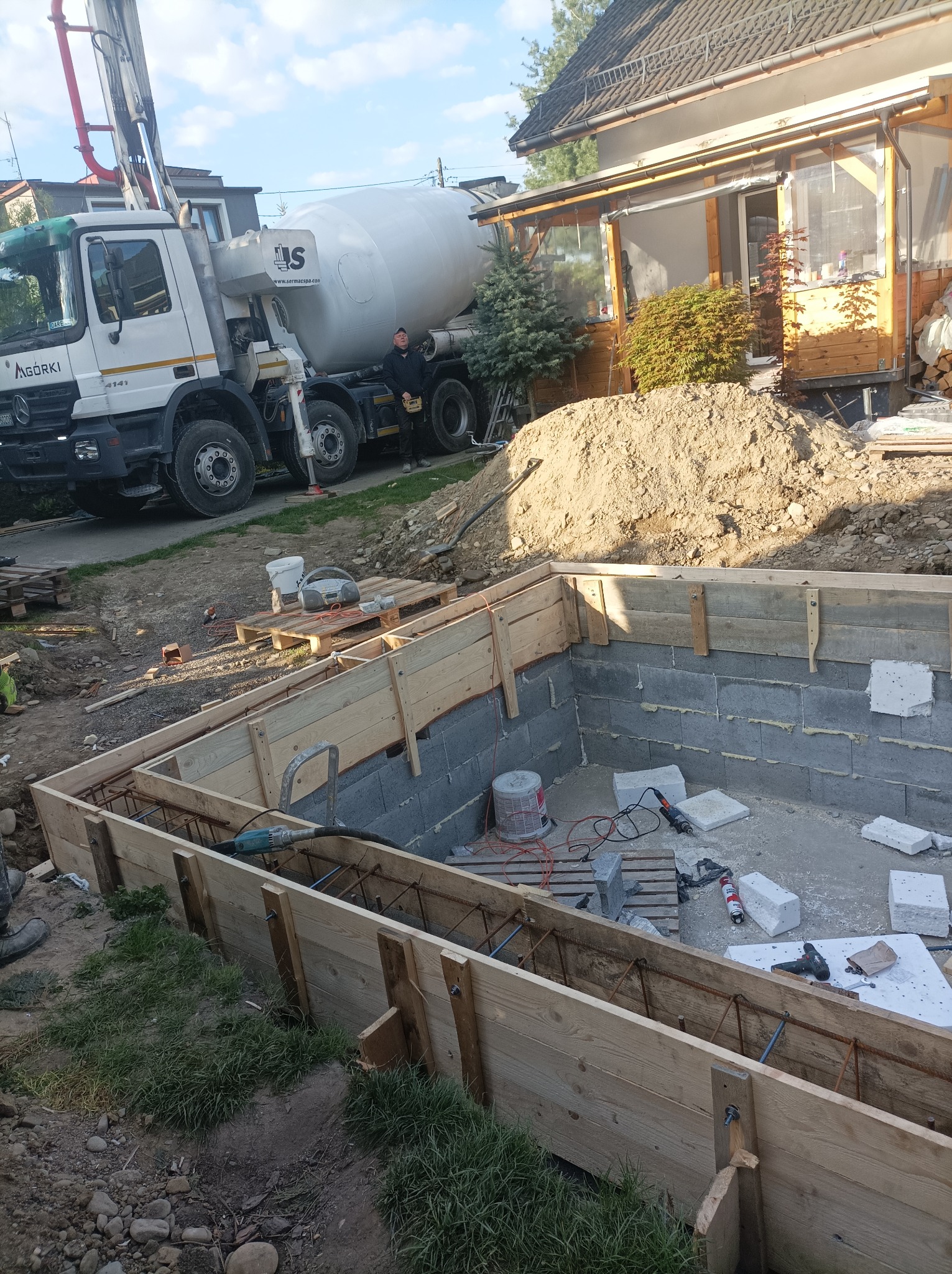 Wykop pod fundamenty z drewnianym szalunkiem i zbrojeniem, widoczny betonowóz i pracownik w tle, narzędzia budowlane rozłożone wewnątrz wykopu.
