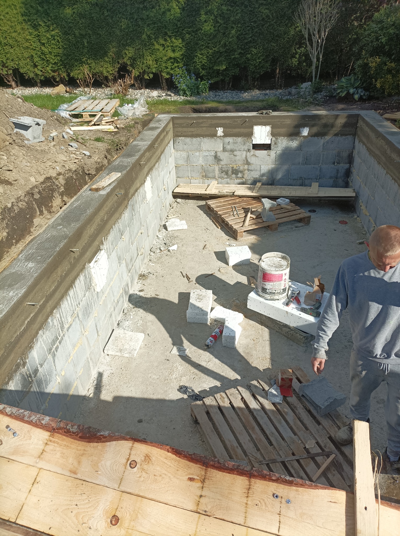 Wykop pod fundamenty z betonową wylewką, widoczne bloczki betonowe, palety drewniane, puszka z klejem i narzędzia, częściowo widoczna sylwetka mężczyzny w szarej bluzie.