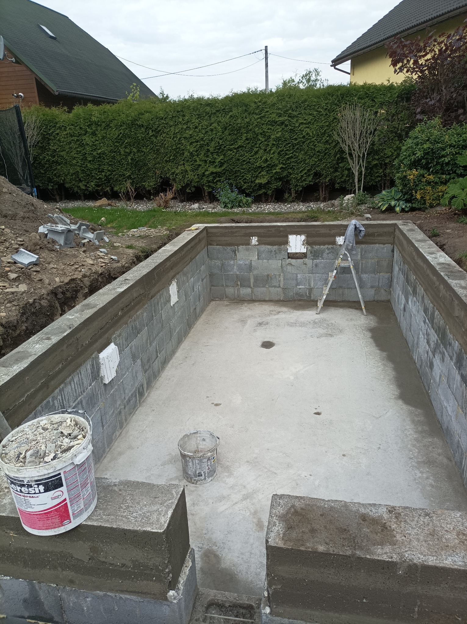 Budowa basenu ogrodowego z szarych bloczków betonowych i wylewką, widoczna drabina oparta o ścianę oraz wiadro z zaprawą klejową 'Ceresit' na pierwszym planie.