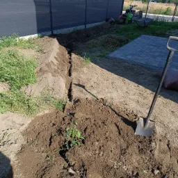Wykopany wąski rów w ziemi wzdłuż ogrodzenia, z łopatą opartą o brzeg, w tle kostka brukowa i sprzęt ogrodniczy.