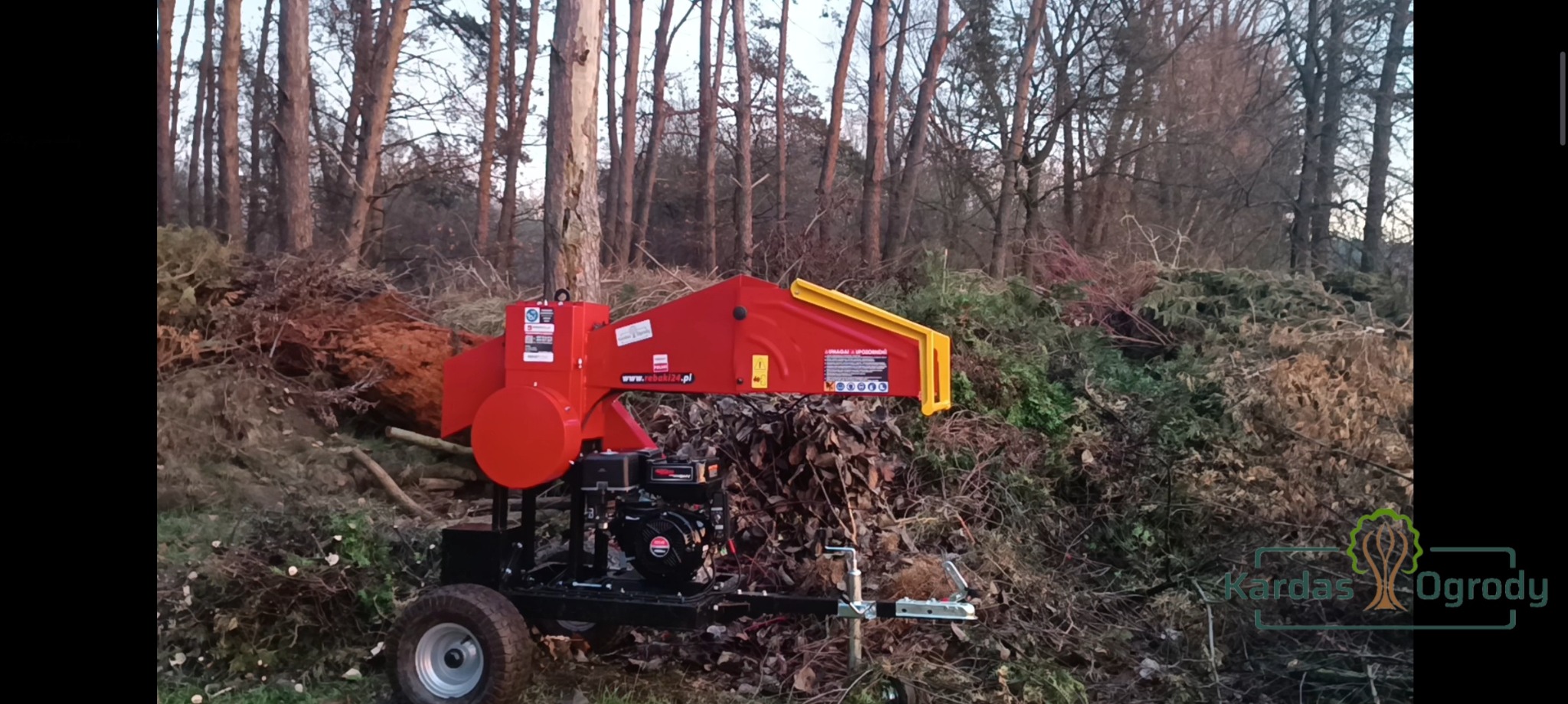 Czerwona rębarka do gałęzi na tle sterty pociętych gałęzi i drzew w lesie, widoczne logo firmy na dole po prawej stronie.