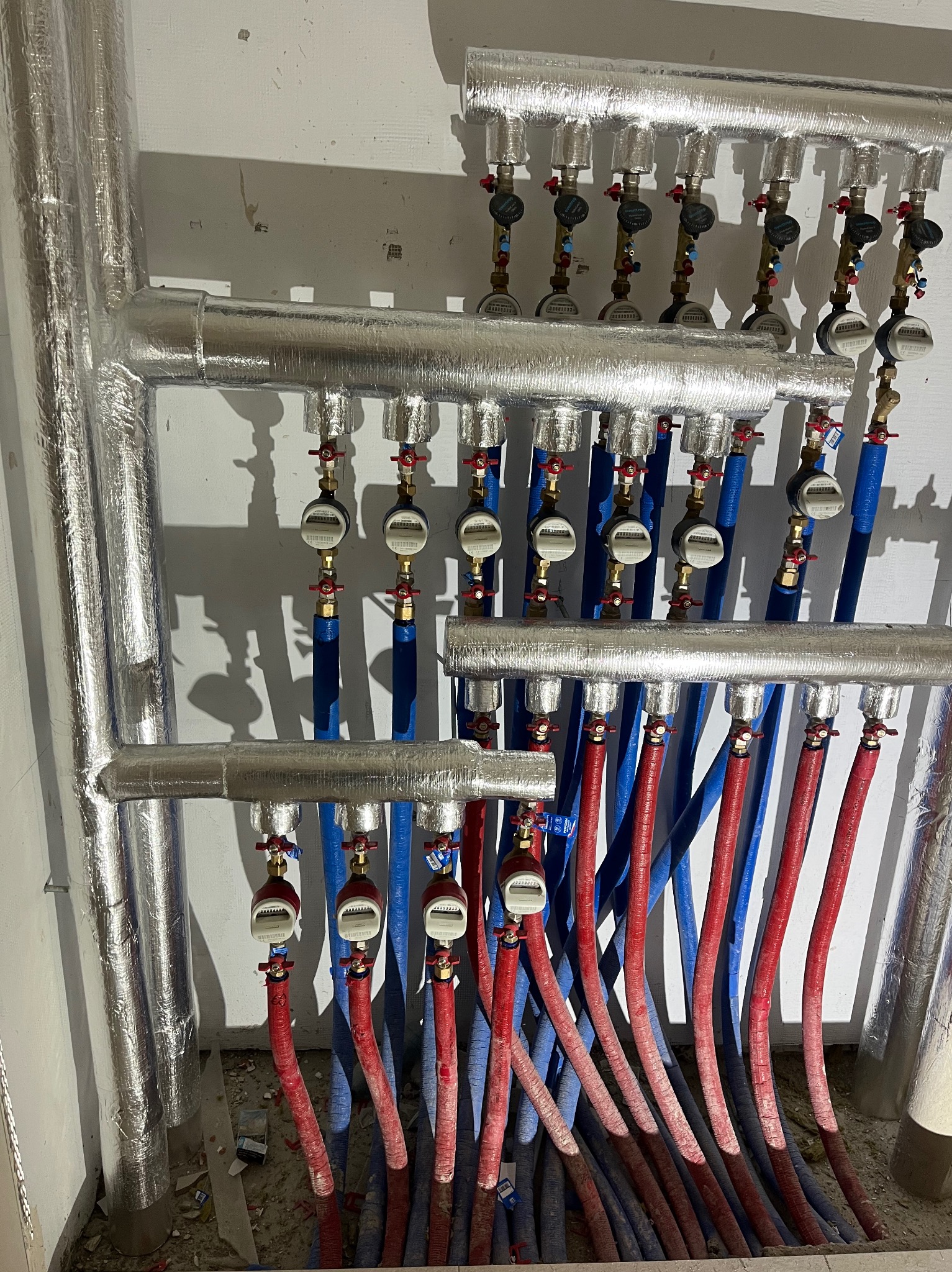 Rozdzielacz CO z licznymi zaworami, miernikami i rurami w kolorach niebieskim i czerwonym, zabezpieczonymi izolacją. Instalacja w surowym wnętrzu.