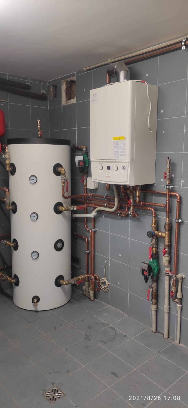Biały piec gazowy i zasobnik ciepłej wody z miedzianą instalacją w szarej kotłowni. Widoczne manometry i zawory. Ukończona instalacja grzewcza.