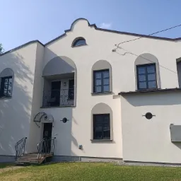 Wykończona elewacja dwupiętrowego domu z balkonem, oknami w ciemnych ramach i ozdobnymi elementami architektonicznymi, widok z poziomu trawnika.