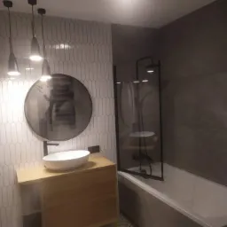 Nowoczesna łazienka z owalnym lustrem, umywalką nablatową na drewnianej szafce, białą wanną z czarnym parawanem i szarą glazurą na ścianach, oświetlona trzema wiszącymi lampami.