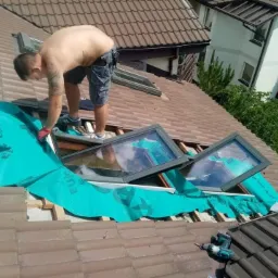 Pracownik bez koszulki w czerwonych rękawicach montuje okna dachowe na brązowym dachu z użyciem zielonej membrany hydroizolacyjnej, w tle widoczny budynek mieszkalny i drzewa.