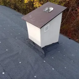 Szczegółowe ujęcie dachu pokrytego papą z widocznym kominem w Warka, otoczonym jesiennymi drzewami.