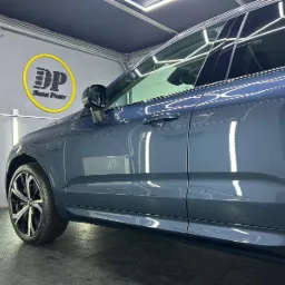 Błyszczący, szary samochód SUV z przyciemnionymi szybami, zaparkowany w dobrze oświetlonym garażu z logo firmy detailerskiej na ścianie.