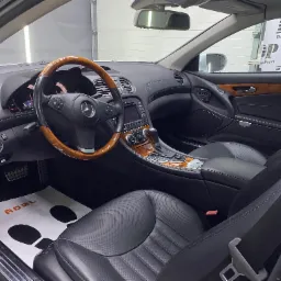 Wnętrze luksusowego samochodu Mercedes SL z drewnianą kierownicą i czarną skórzaną tapicerką po zabiegach pielęgnacyjnych, widoczna mata ochronna na podłodze z logo ADBL oraz logo DarkPoint...