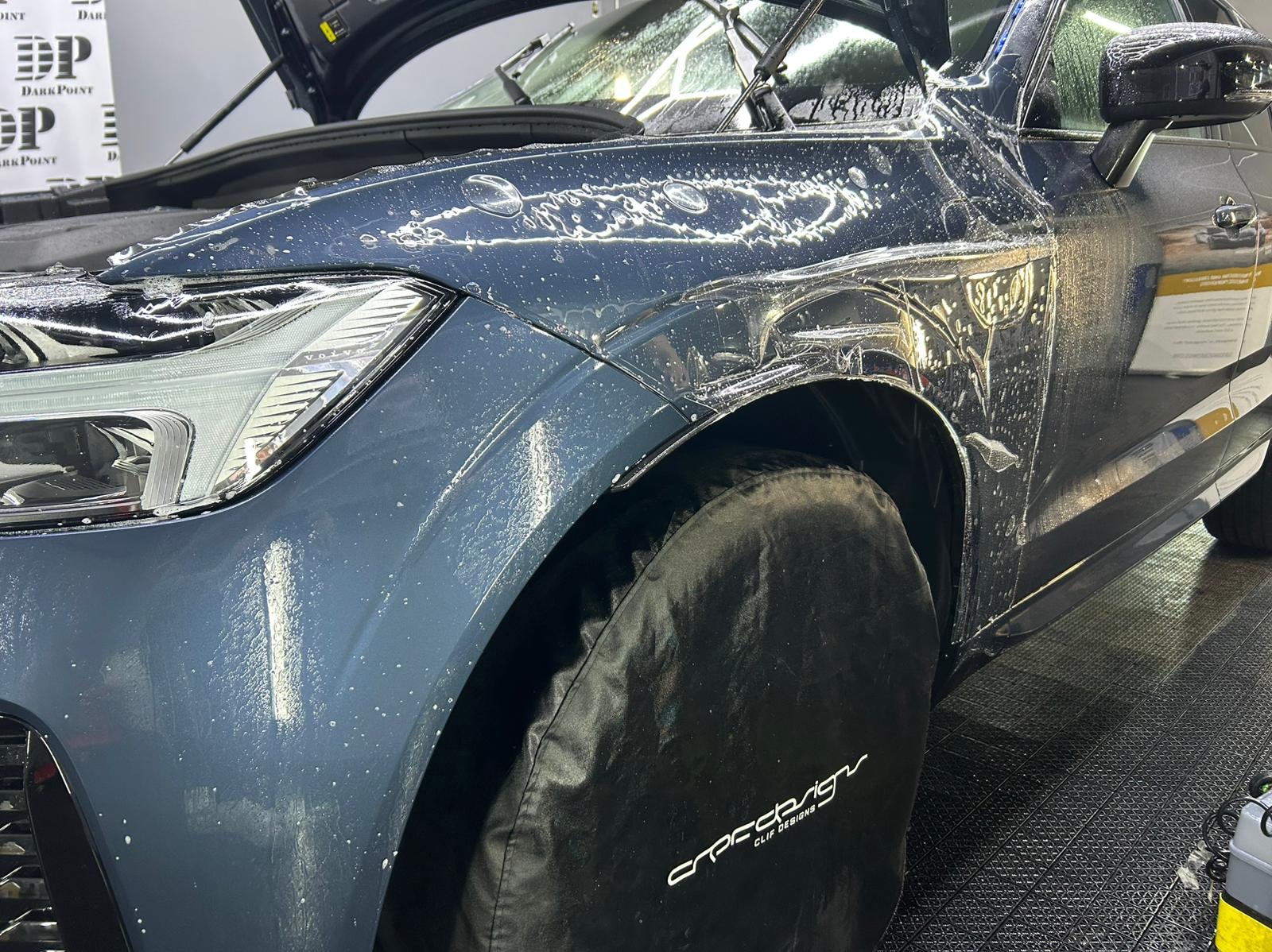 Niebieski samochód marki Volvo w trakcie aplikacji folii ochronnej na przedni błotnik i reflektor, z zakrytym kołem i widocznymi pęcherzykami wody pod folią.