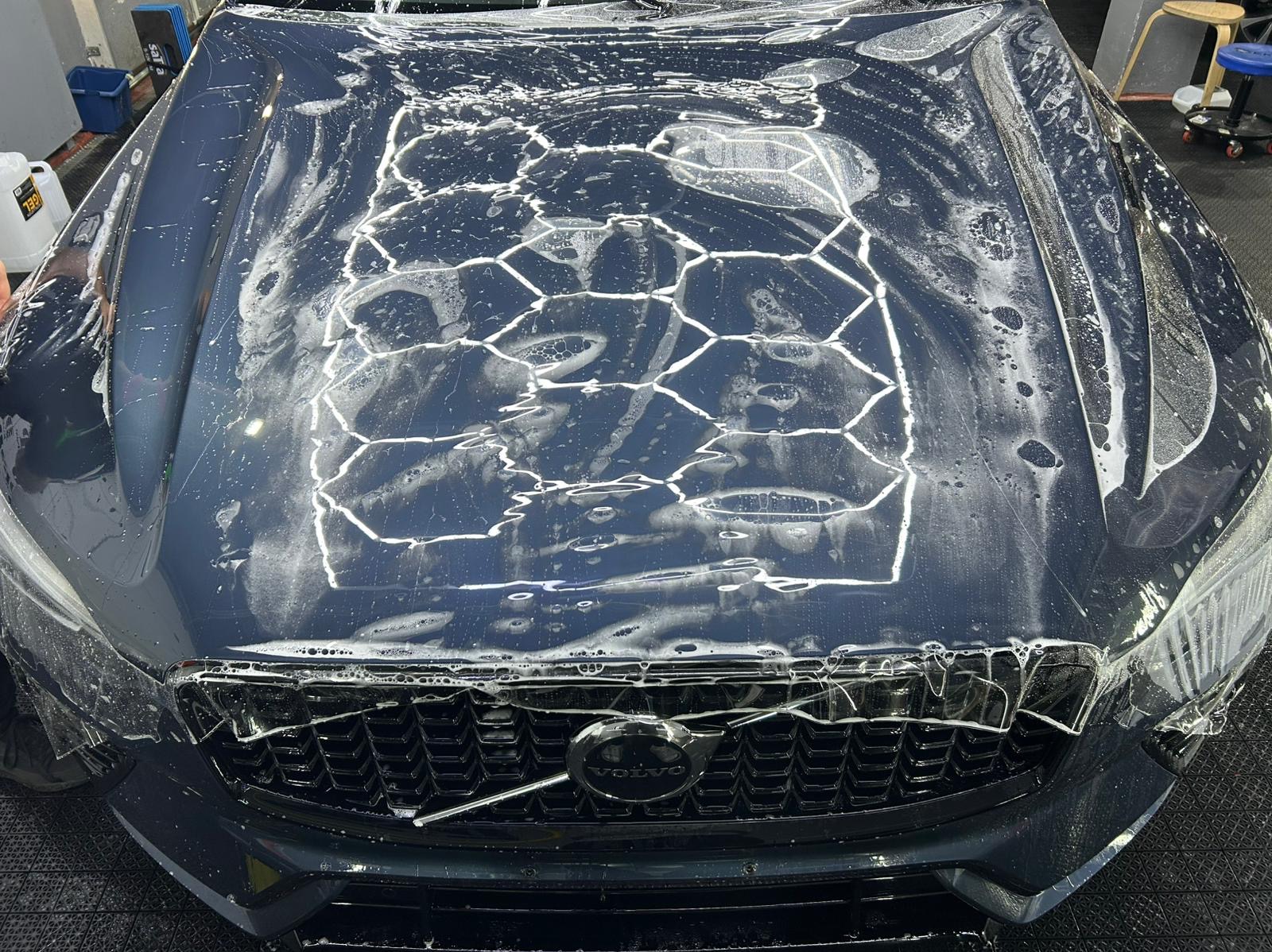 Proces aplikacji folii ochronnej na maskę samochodu Volvo, widoczne pęcherzyki powietrza i woda, zbliżenie na grill i logo.